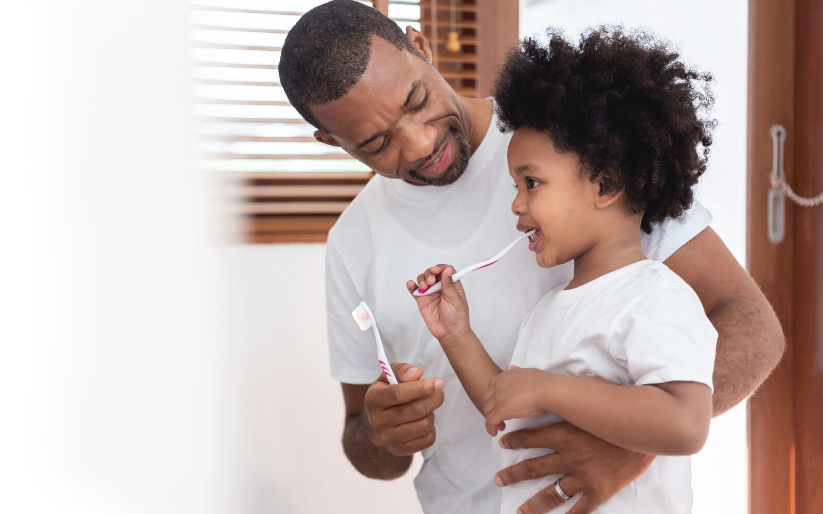 Happy African American Family, Dad teaching his son in white shirts brushing teeth in bathroom together. Father and kid boy in curly hair enjoying with dental hygiene at home in morning